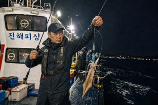 Edit the source image into one polished final result.
Primary intent: 원본 인물과 야간 선상낚시 장면은 유지한다. 손에 든 채비 부분을 실제 한치 오모리그처럼 삼각도래에서 긴 목줄의 에기가 옆으로 분기되고 봉돌은 아래쪽으로 늘어지는 구조로 정확하고 자연스럽게 수정한다. 낚싯줄의 연결점, 장력, 비례와 금속 부품을 사실적으로 표현하고 한치의 포획 상태, 조명, 원근, 물방울은 장면에 일관되게 유지한다.
Conversation context to respect: User concern to address: 그래 / Resolved edit decision: 실제 한치 오모리그와 정확히 맞추려면 먼저 웹에서 표준 채비 사진을 확인해 연결 구조를 기준으로 삼는 게 좋겠습니다. 참고 이미지를 찾아본 뒤 자연스럽게 수정할까요
Keep the overall composition, identity, and scene continuity intact.
Apply the change mainly to the requested subject and the immediately adjacent area.
Do not introduce unrelated new elements or broad scene changes.
Make the result look intentional, realistic, and production-ready.