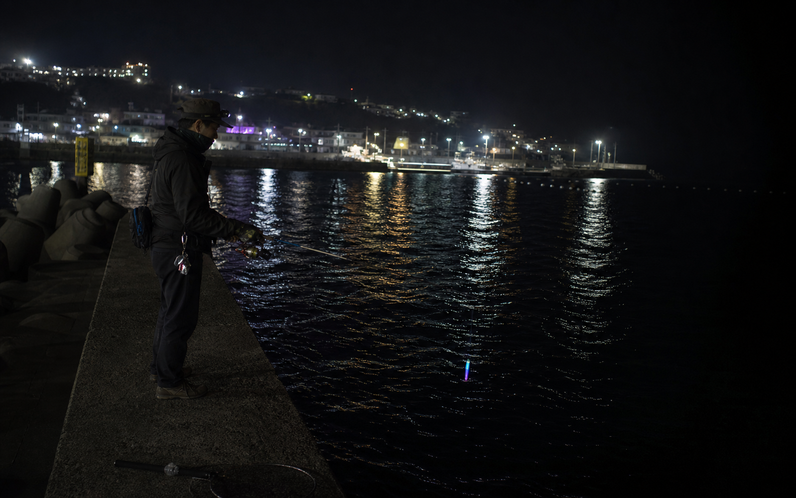 Night eging scene on a Korean breakwater, one angler targeting the boundary between harbor lights and dark water, reflective sea surface, subtle squid lure glow, realistic low-light photography, cinematic but natural
