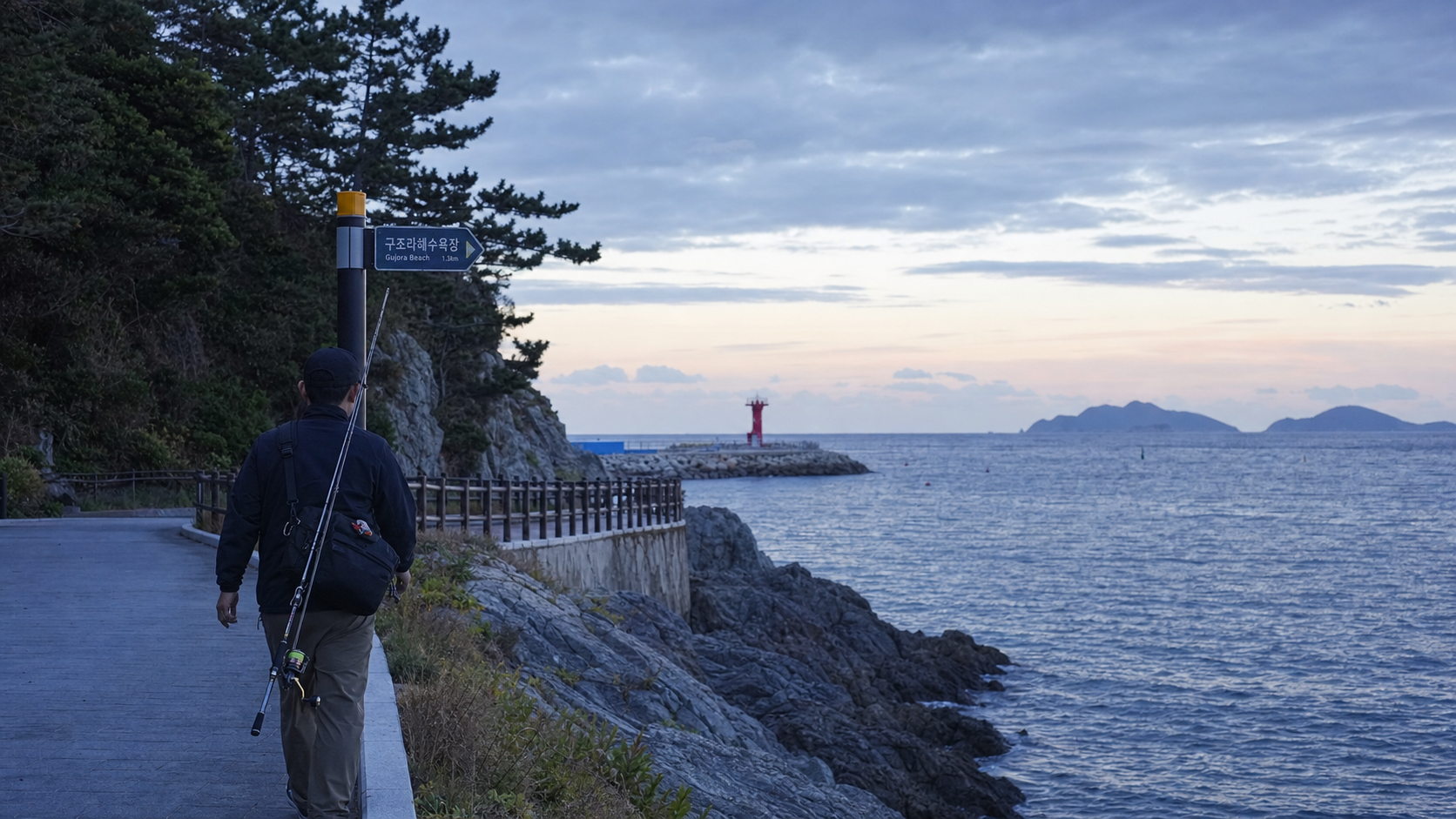 (Documentary coastal fishing scene at Geoje south coast, beginner angler walking on a clean seaside boardwalk toward a breakwater at dawn, compact eging rod, sling bag, calm but open sea, subtle lighthouse in distance, realistic Korean shoreline texture, 35mm lens, natural blue hour light, editorial travel-fishing mood --ar 16:9) 초행자가 거제 남부권 도보 에깅에 들어가는 첫 장면. 산책로 접근성과 열린 시야가 동시에 느껴지도록 구성합니다.