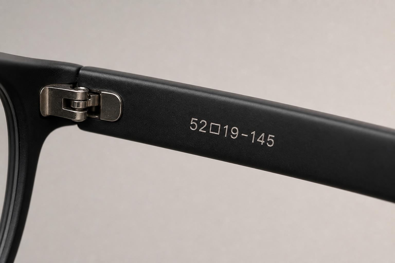 close-up editorial product photo of eyeglasses temple arm with engraved size numbers like 52□19-145, neutral soft lighting, sharp macro focus, matte black frame with subtle metallic hinge, realistic studio background