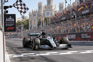 A realistic Formula 1 race scene at a modern Madrid street circuit, Mercedes car crossing the finish line under bright afternoon light, grandstands full of fans, crisp motorsport photography style