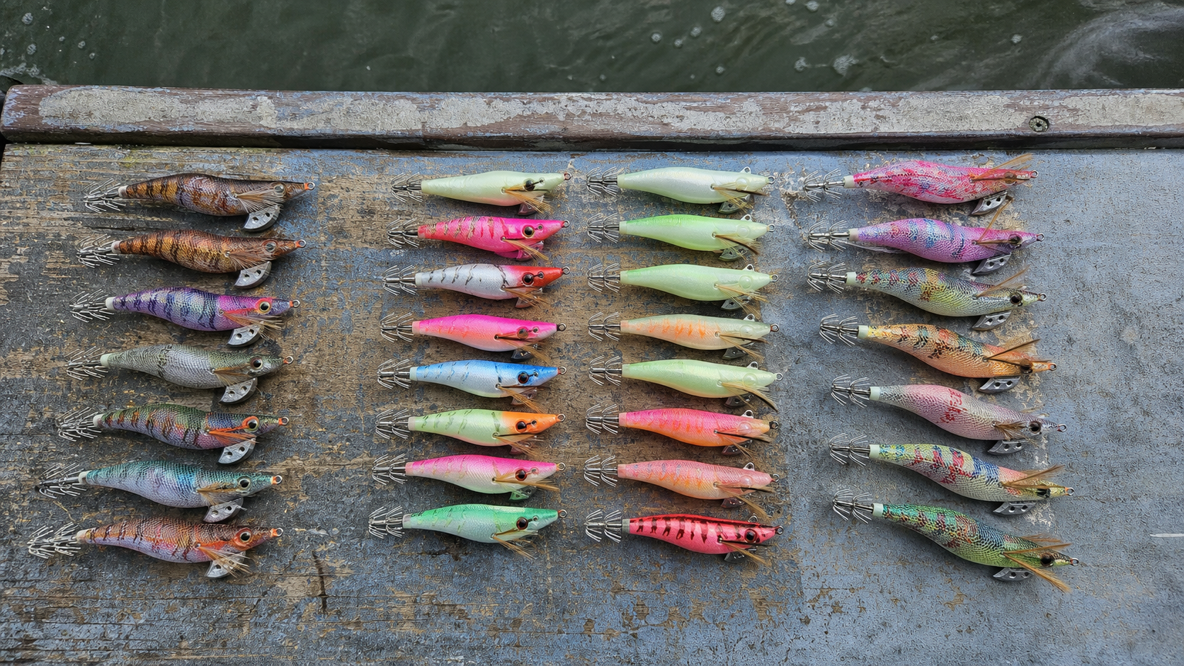 (Overhead editorial product photo of multiple cuttlefish egis arranged by sink rate and color on a weathered boat deck, soft overcast daylight, realistic saltwater texture, polarized reflections controlled, 50mm lens, ultra-detailed tackle catalog style --ar 16:9) 갑오징어 에기를 호수, 발광 타입, 컬러별로 정리한 실전 준비 컷. 장비 비교형 글의 첫 이미지로 적합합니다.